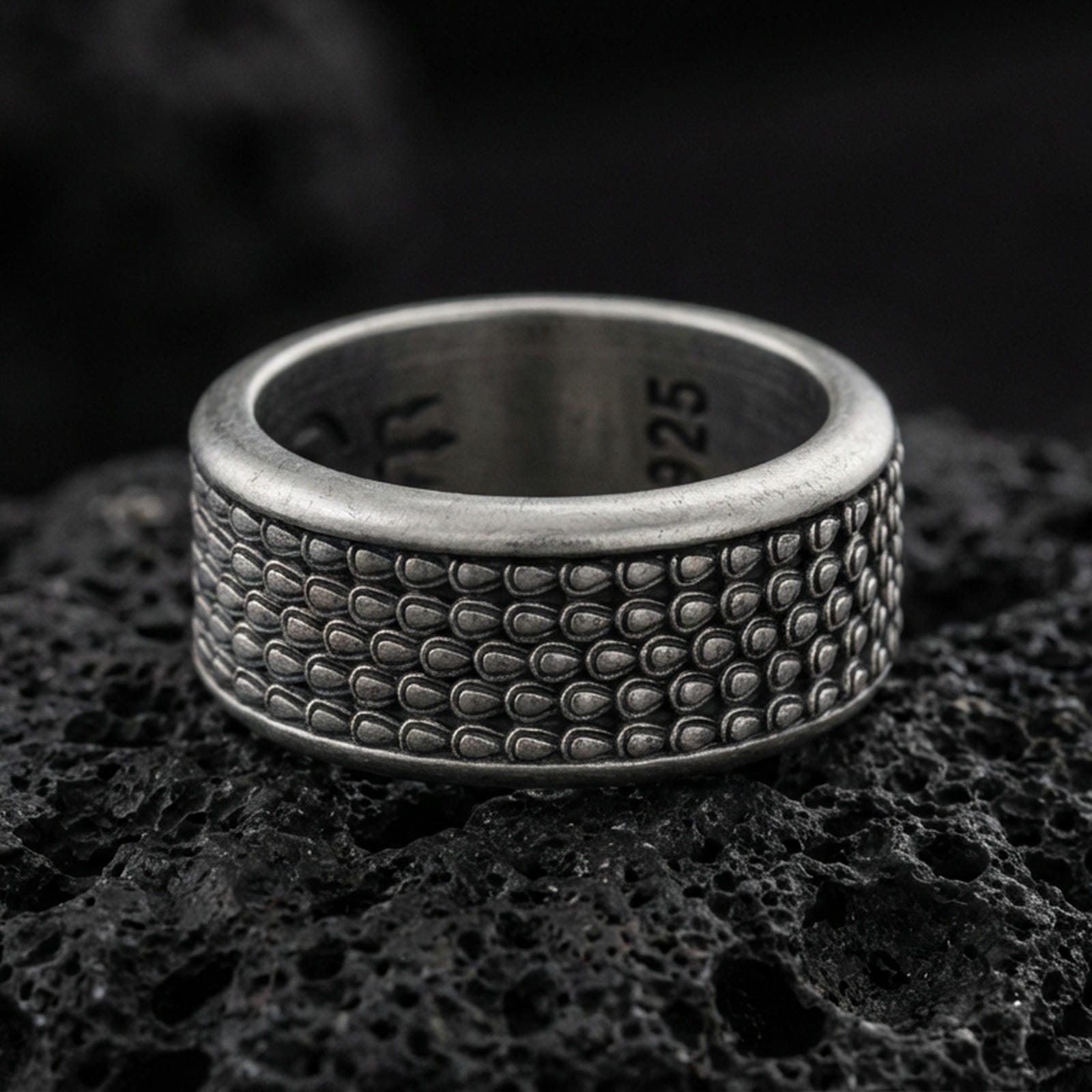 Oxidized sterling silver scale texture band ring on dark volcanic rock, repeating teardrop scale pattern with deep contrast, premium macro product shot.