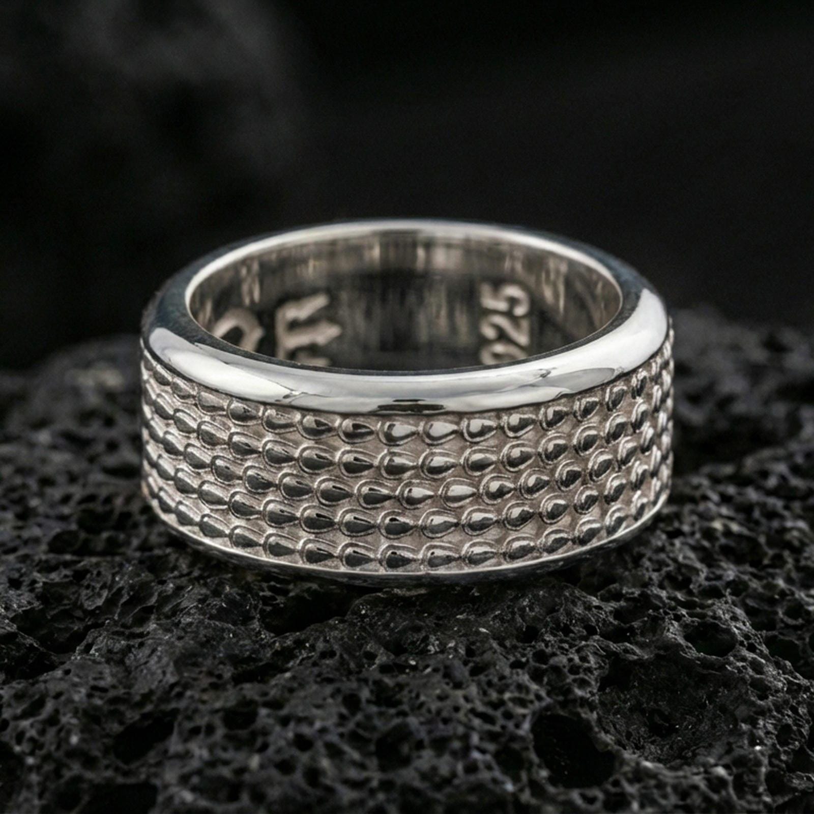 Polished silver textured scale band ring on black volcanic stone, crisp repeating scale pattern and smooth comfort edges, centered close-up studio lighting.