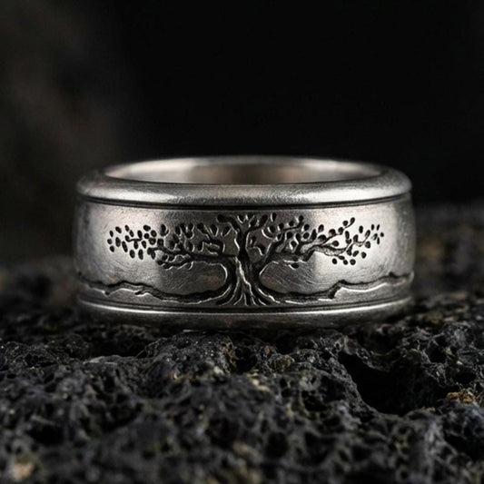 Oxidized sterling silver Yggdrasil spinner band ring on dark volcanic rock, engraved tree of life on rotating center with mountain line detail, premium macro product shot.