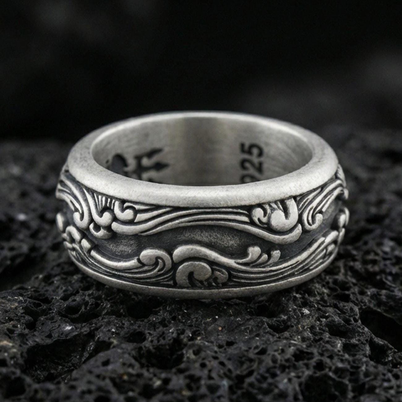 Oxidized sterling silver scrollwave spinner band ring on dark volcanic rock, flowing wave filigree engraving on rotating center, premium macro product shot.