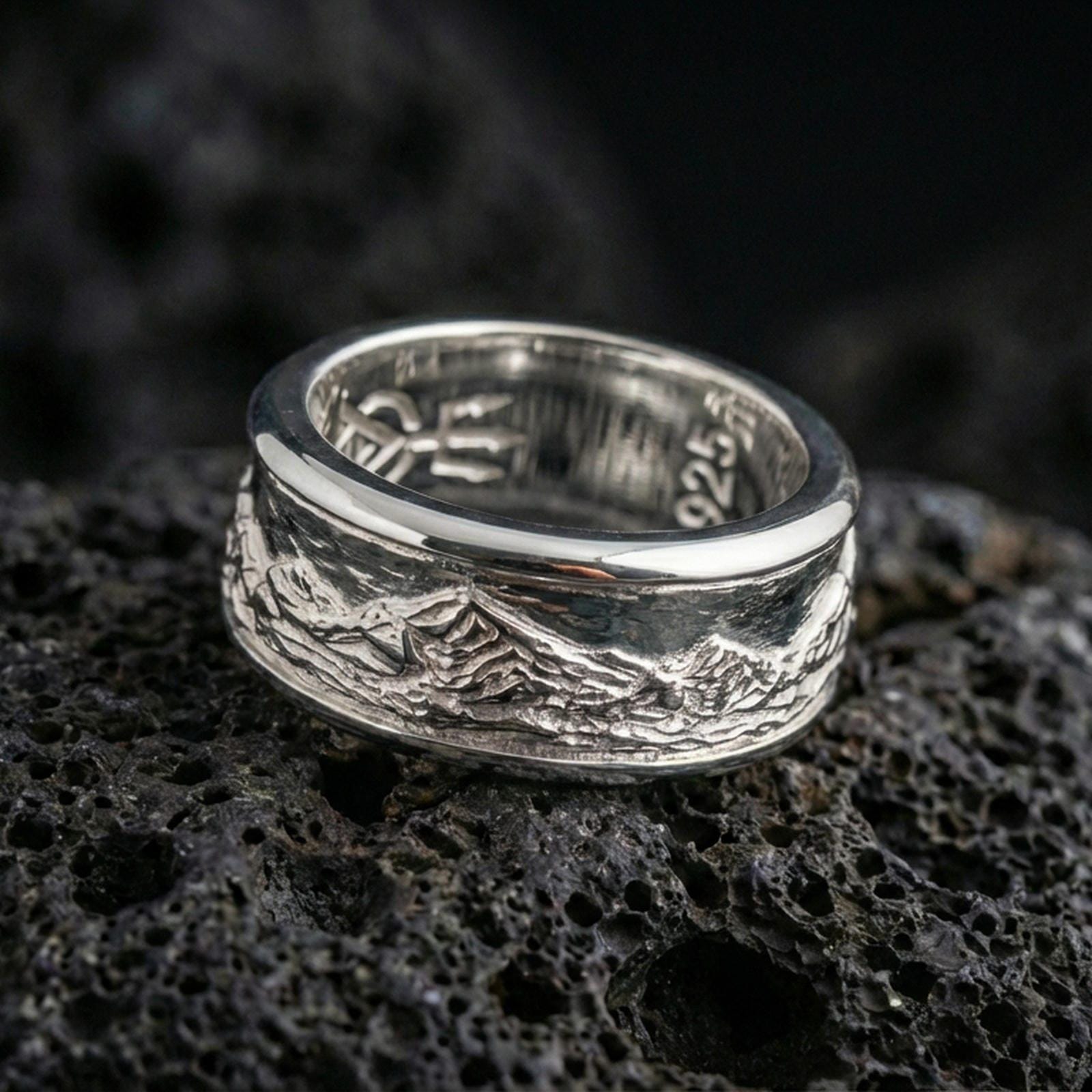Polished silver mountain landscape band ring on black volcanic stone, crisp peak skyline engraving with brushed finish, centered close-up studio lighting.