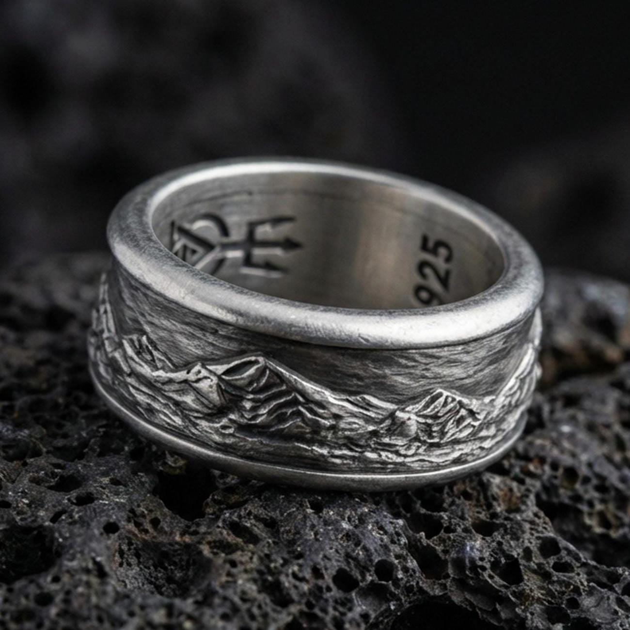Oxidized sterling silver mountain range band ring on dark volcanic rock, rugged alpine skyline engraving with deep contrast, premium macro product shot.