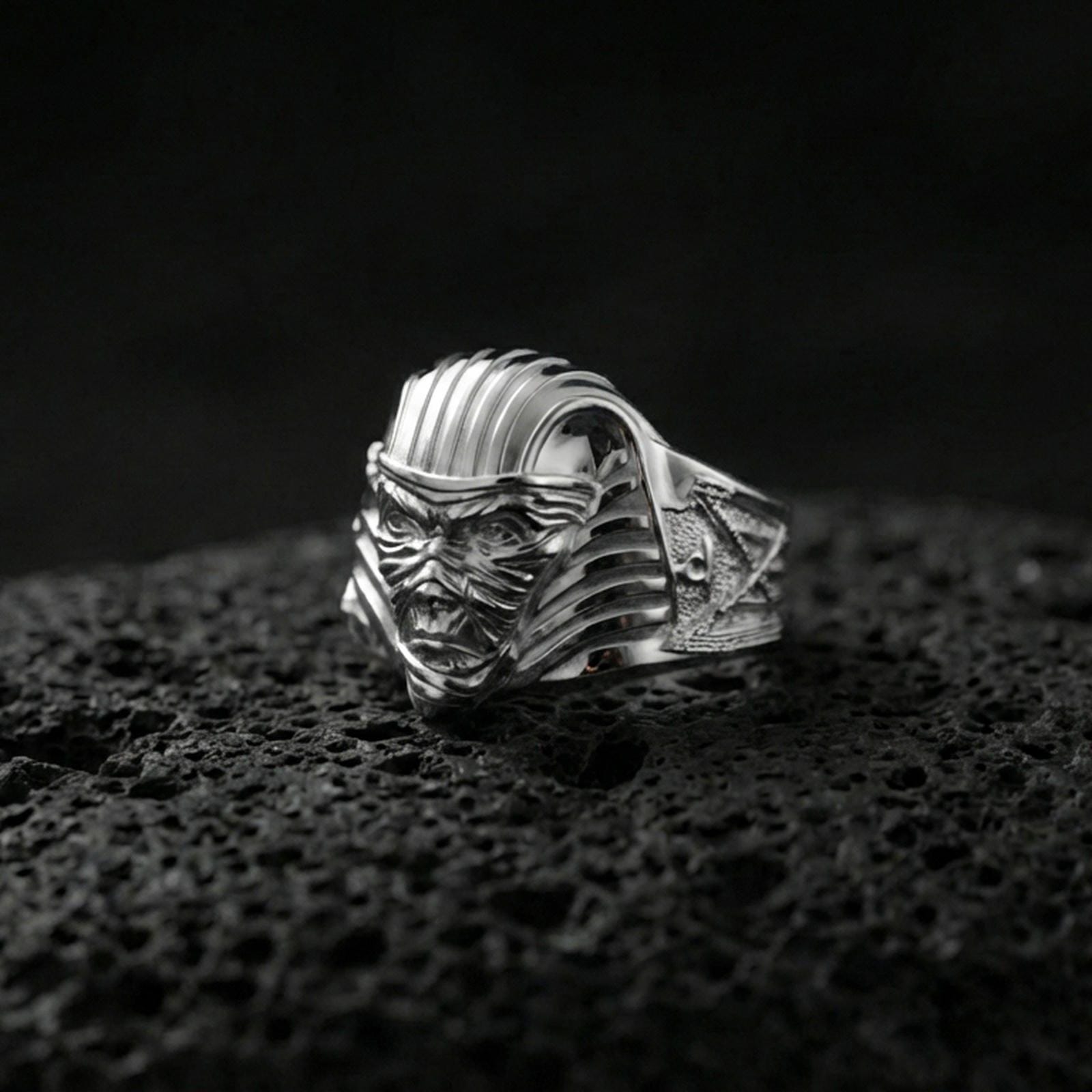 Sterling silver Egyptian mummy mask statement ring in a polished silver finish, sculpted monster face with striped pharaoh-style headpiece, close-up on volcanic stone