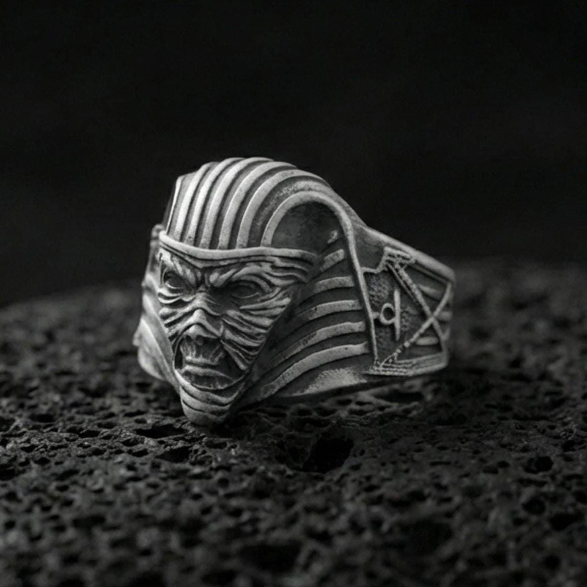Sterling silver mummy pharaoh mask ring with an oxidized finish, horror-inspired Egyptian face design with ribbed headdress detail, macro on black volcanic rock