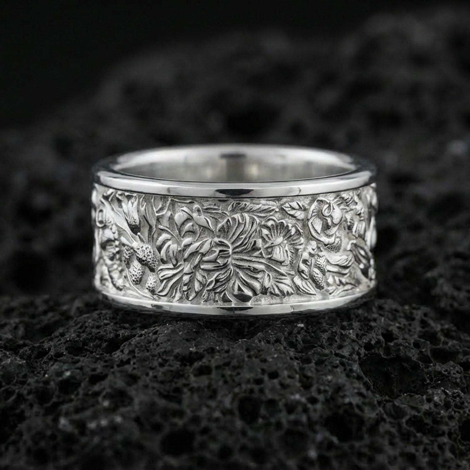Polished silver nature wildlife engraved band ring on black volcanic stone, crisp botanical relief with detailed jungle scene texture, centered close-up studio lighting.