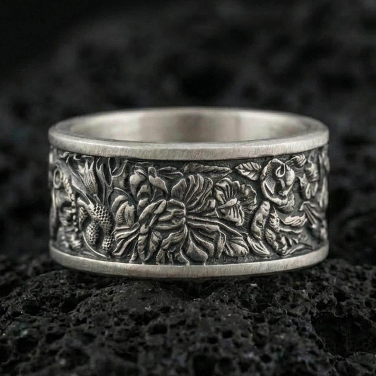 Oxidized sterling silver jungle wildlife band ring on dark volcanic rock, high-relief leaves and exotic animal engraving, premium macro product shot.