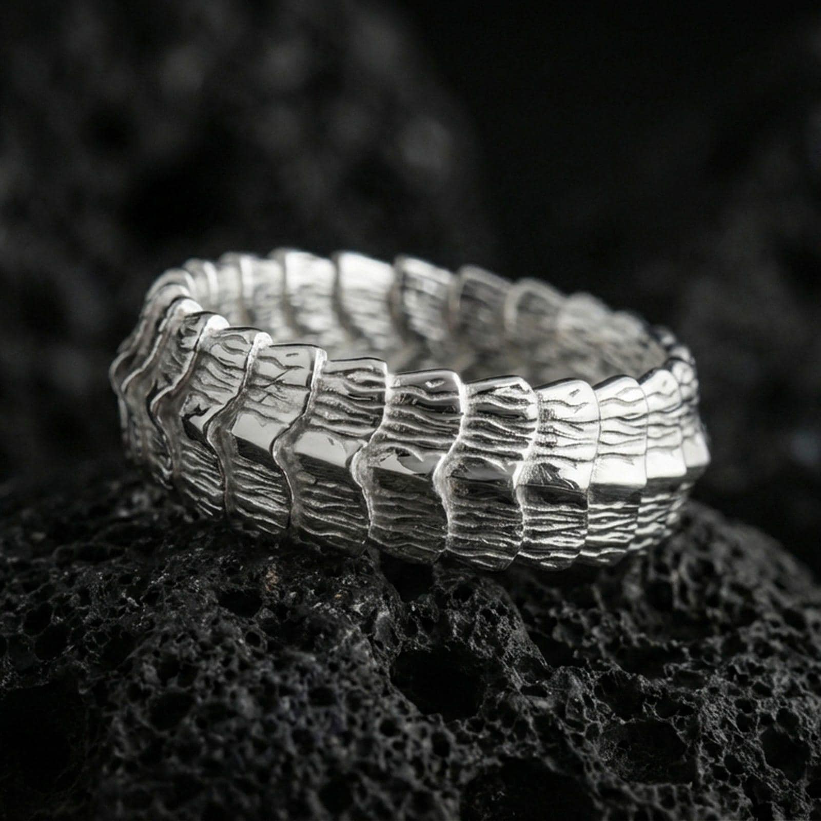 Polished silver gothic dragon scale band ring on black volcanic stone, crisp layered scale ridges with sculpted organic texture, centered close-up studio lighting.
