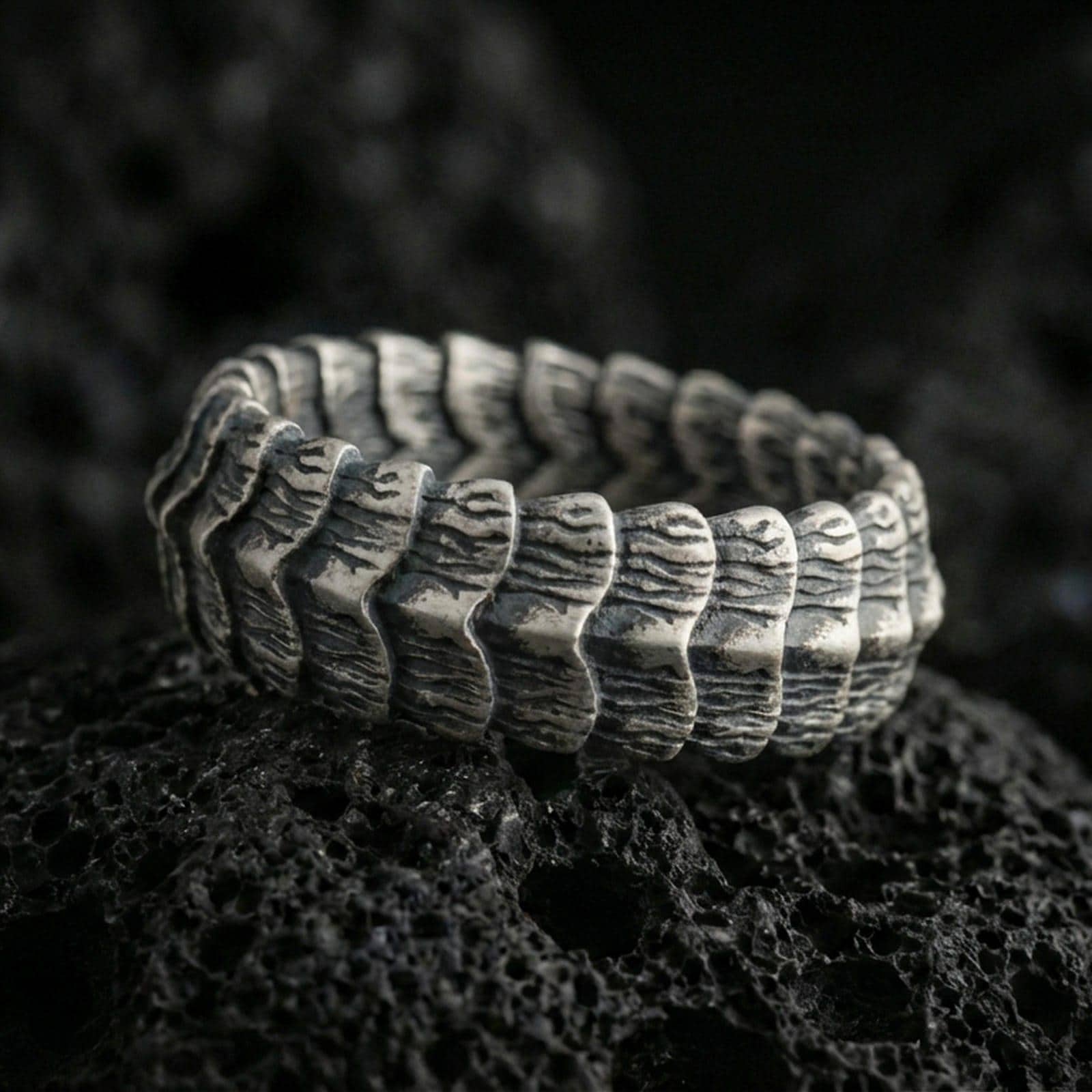 Oxidized sterling silver dragon scale band ring on dark volcanic rock, rugged segmented ridge texture with deep contrast, premium macro product shot.