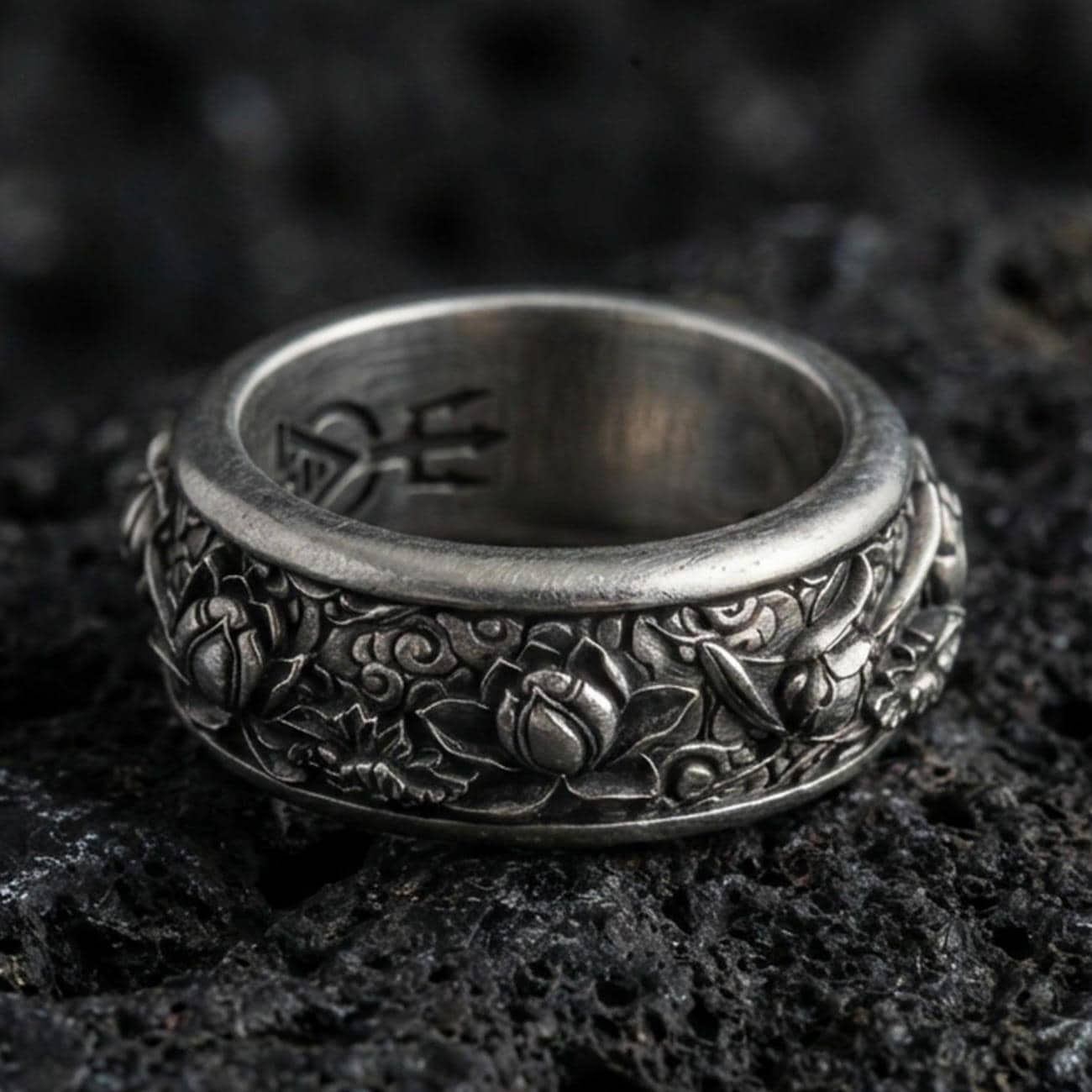 Oxidized sterling silver lotus spinner band ring on dark volcanic rock, raised lotus flowers and scrollwork on rotating center, premium macro product shot.