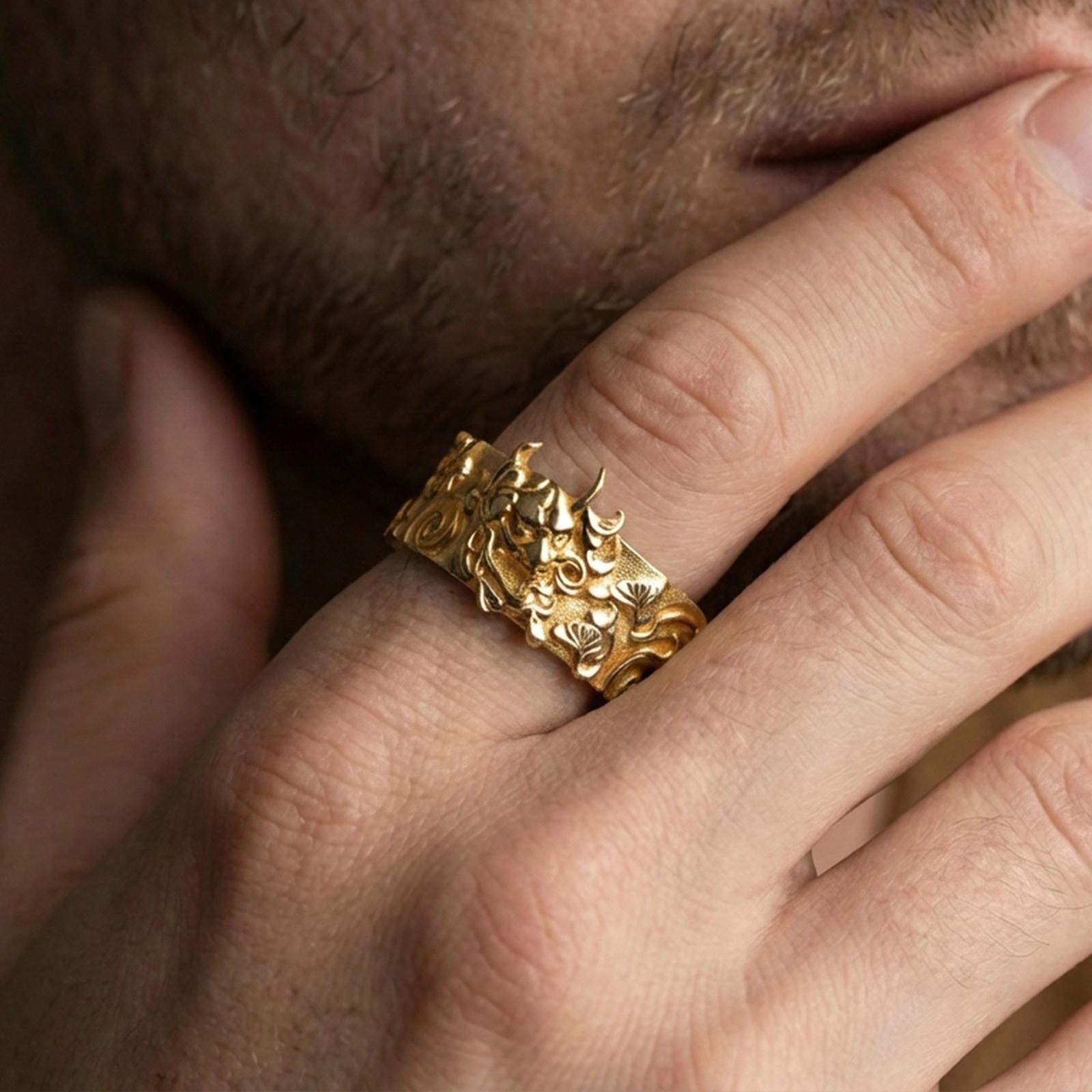 Gold plated oni mask band ring worn on finger, detailed Japanese demon face relief with cloud motifs, premium lifestyle jewelry photo.