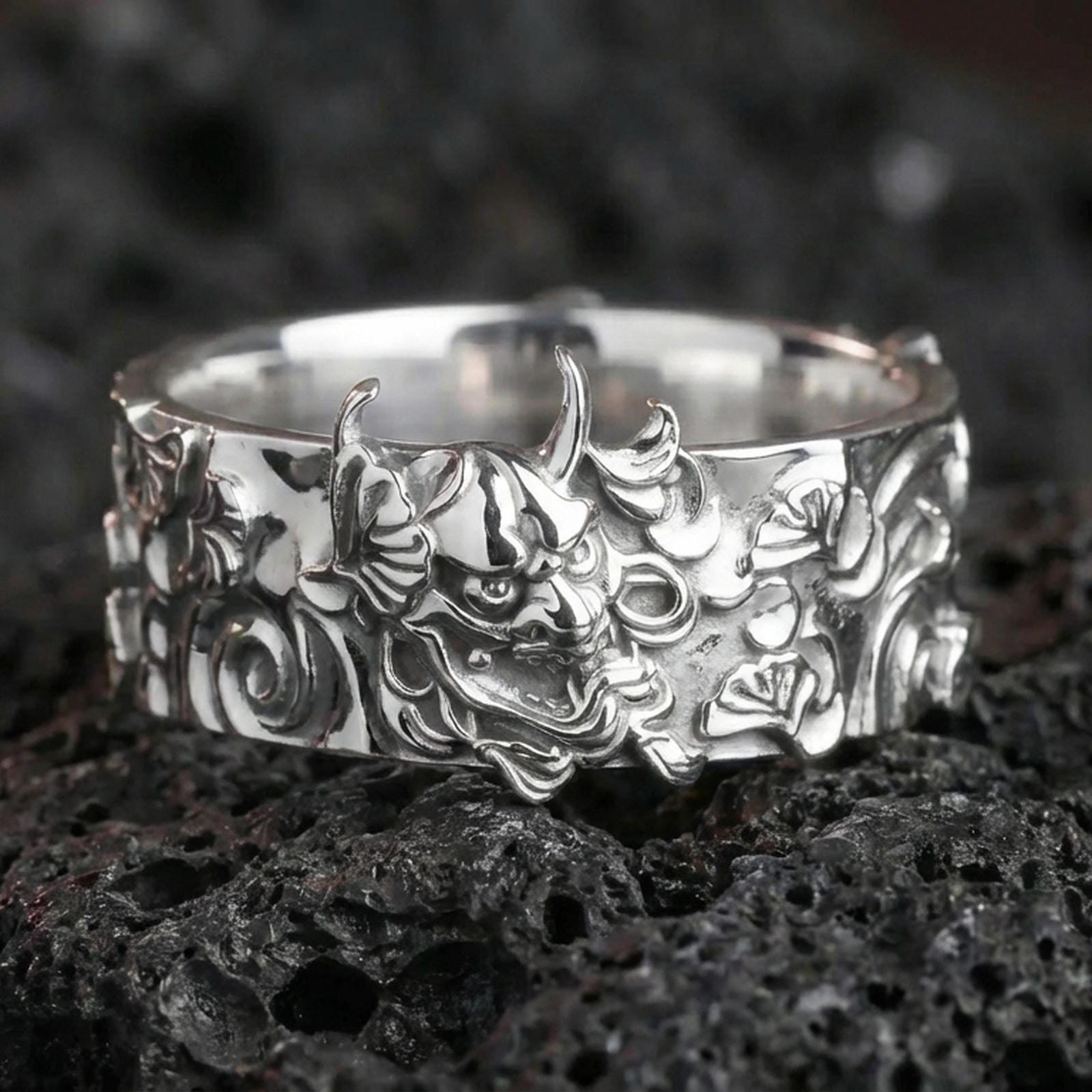 Polished silver oni mask statement ring on black volcanic stone, crisp Japanese demon relief with bold sculpted details, centered close-up studio lighting.