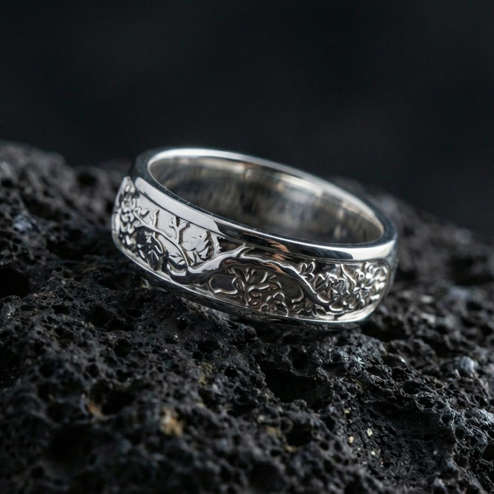 Polished silver nature tree roots band ring on black volcanic stone, crisp branch relief with leaf details and smooth comfort edges, centered close-up studio lighting.