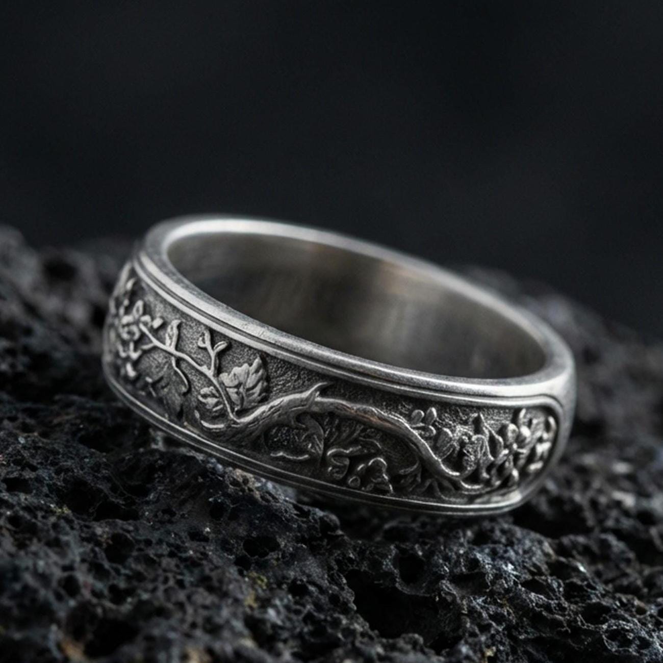 Oxidized sterling silver tree branch band ring on dark volcanic rock, detailed root and leaf vine engraving with woodland texture, premium macro product shot.