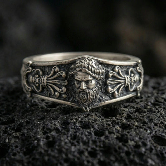 Oxidized sterling silver Zeus portrait band ring on dark volcanic rock, high-relief bearded god face with ornate classical scroll panels, premium macro product shot.