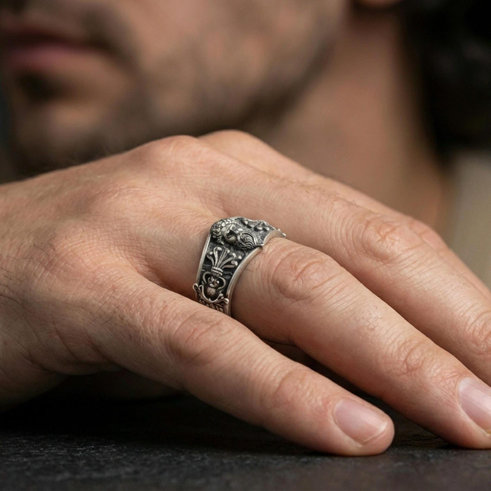 Oxidized sterling silver Zeus portrait band worn on finger, bearded god relief and ornate side motif detail, natural lifestyle hand shot with shallow depth of field.