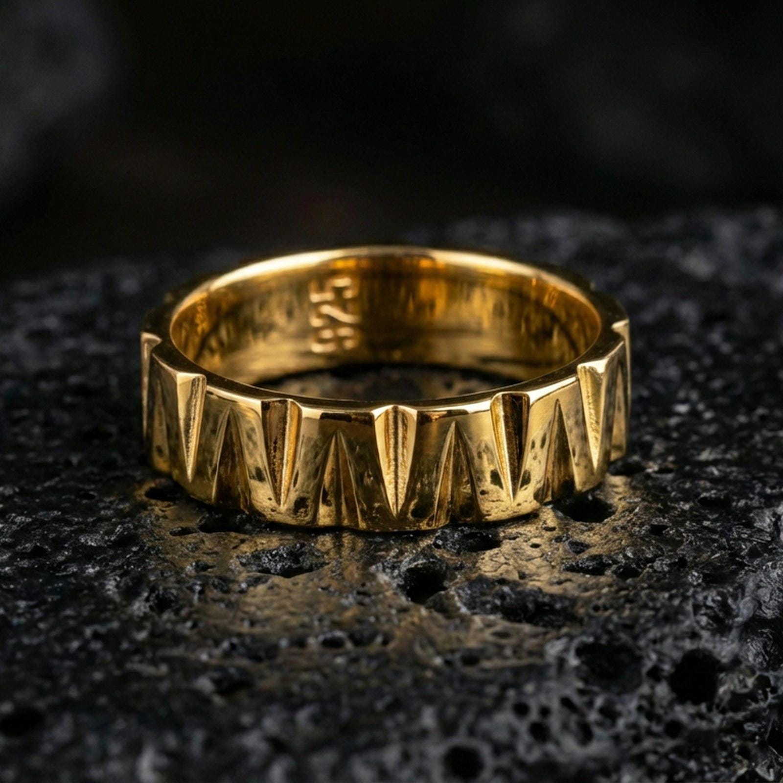 Gold plated faceted chevron band ring on volcanic rock, sharp triangular groove pattern with high-contrast depth, luxury studio macro shot.