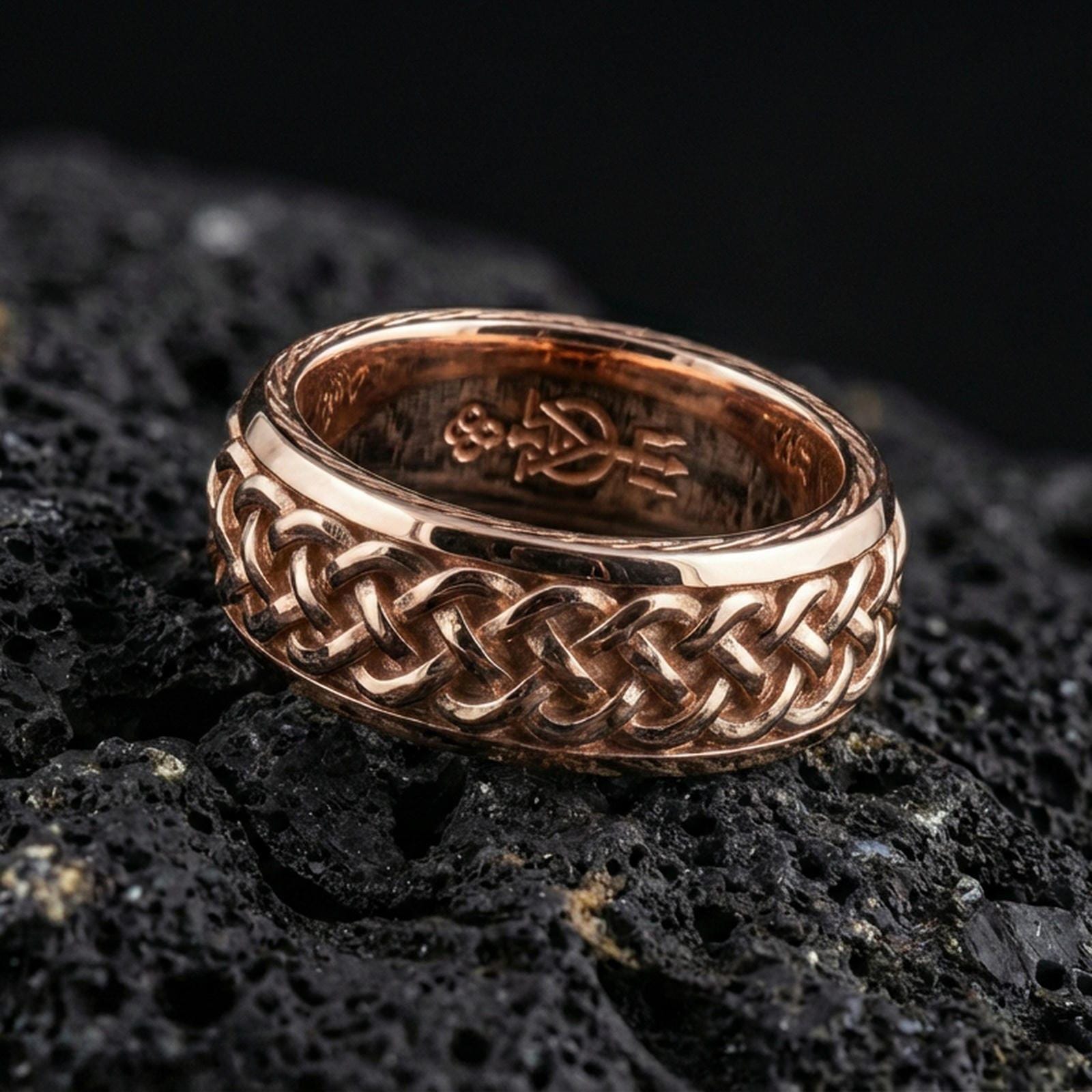 Rose gold plated braided Celtic knot band on black volcanic stone, intricate interlace pattern with warm reflective highlights, refined product close-up.