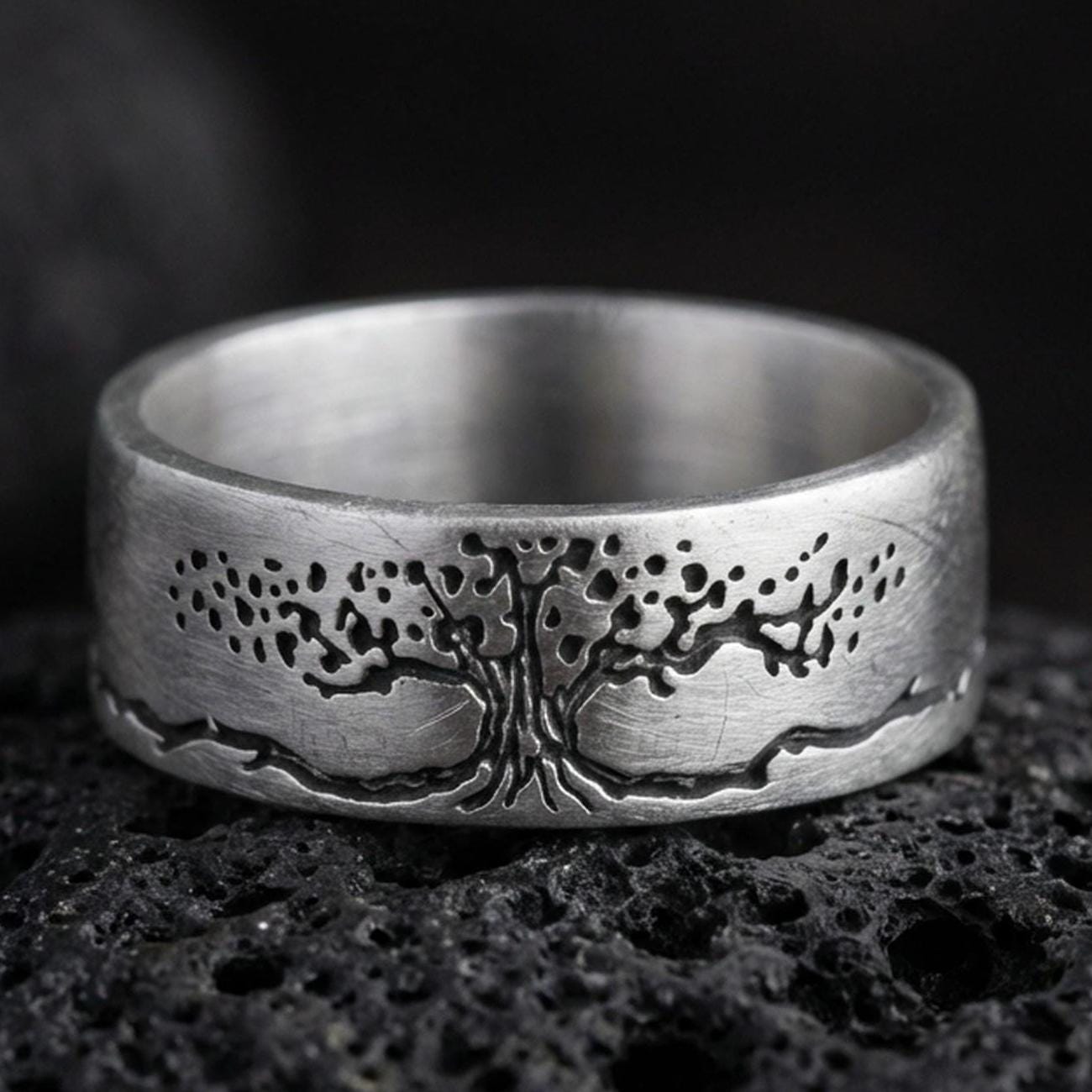 Oxidized sterling silver Tree of Life band ring on black volcanic stone, darkened engraved branches and roots for strong contrast, macro product photo.