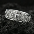 Load image into Gallery viewer, Polished silver gothic skull mechanical band ring on volcanic rock, bright reflective finish emphasizing the raised skull, flowing filigree lines, and crisp gear texture, premium close-up.
