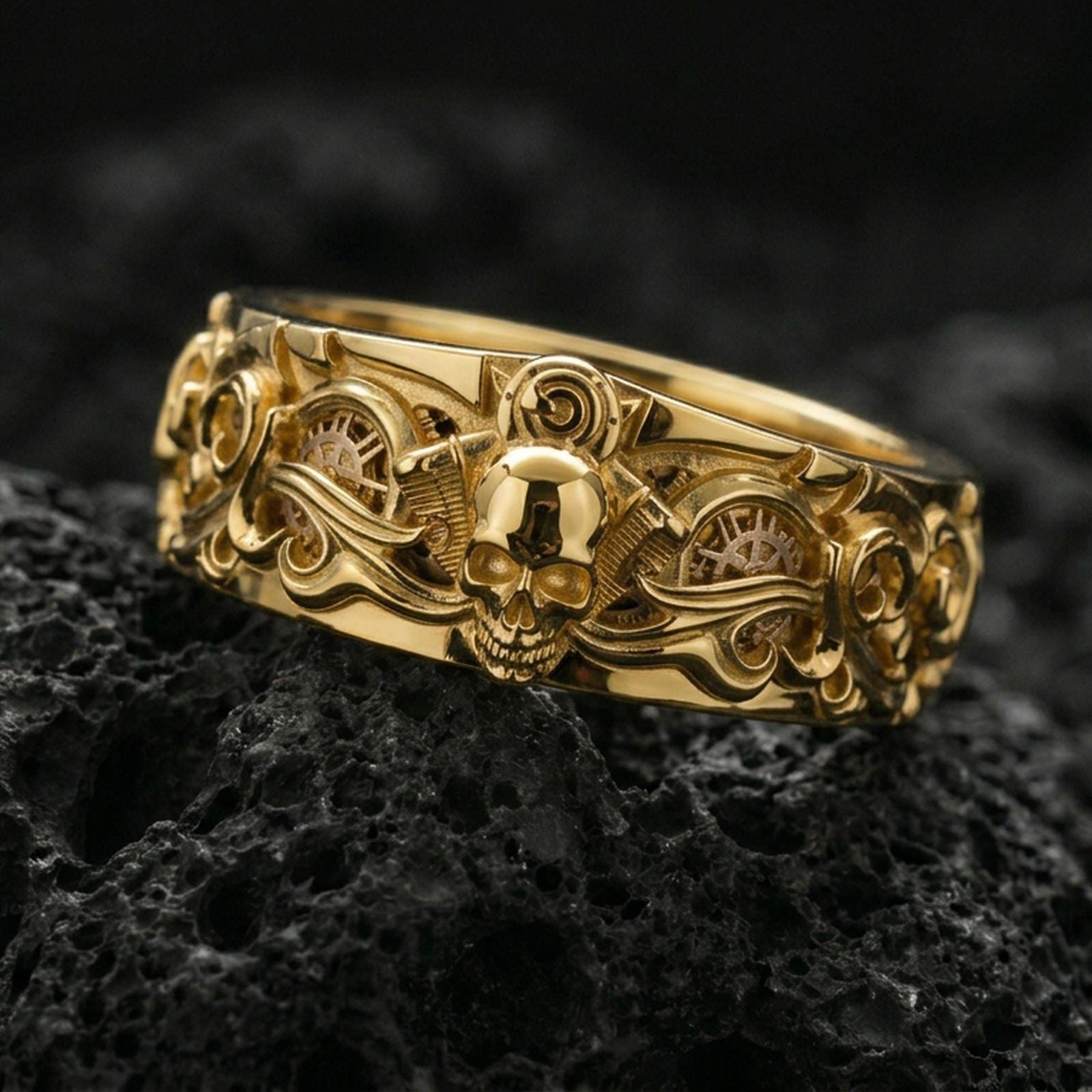 Gold plated skull and gears statement band ring on black volcanic stone, warm shine defining the sculpted skull face and mechanical accents with strong depth, studio macro.