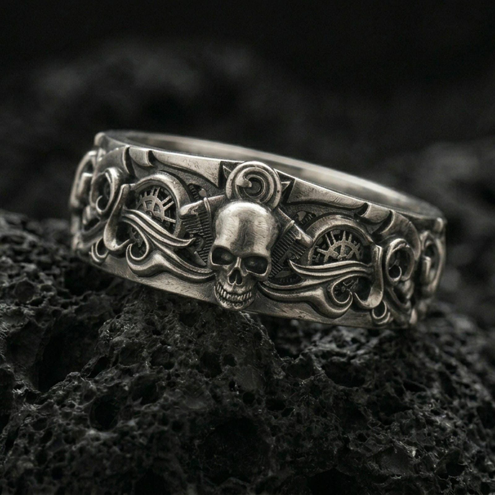 Oxidized sterling silver gothic skull gear band ring on black volcanic stone, darkened relief highlighting the central skull, side scrollwork, and industrial gear details, macro product photo.
