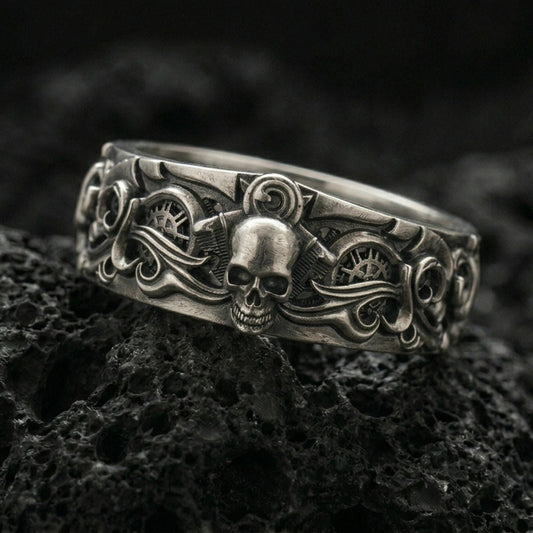 Oxidized sterling silver gothic skull gear band ring on black volcanic stone, darkened relief highlighting the central skull, side scrollwork, and industrial gear details, macro product photo.