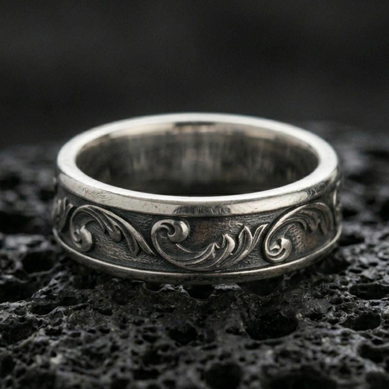 Oxidized sterling silver floral scroll band ring on black volcanic stone, vintage darkened grooves emphasizing the minimalist engraved vine pattern.