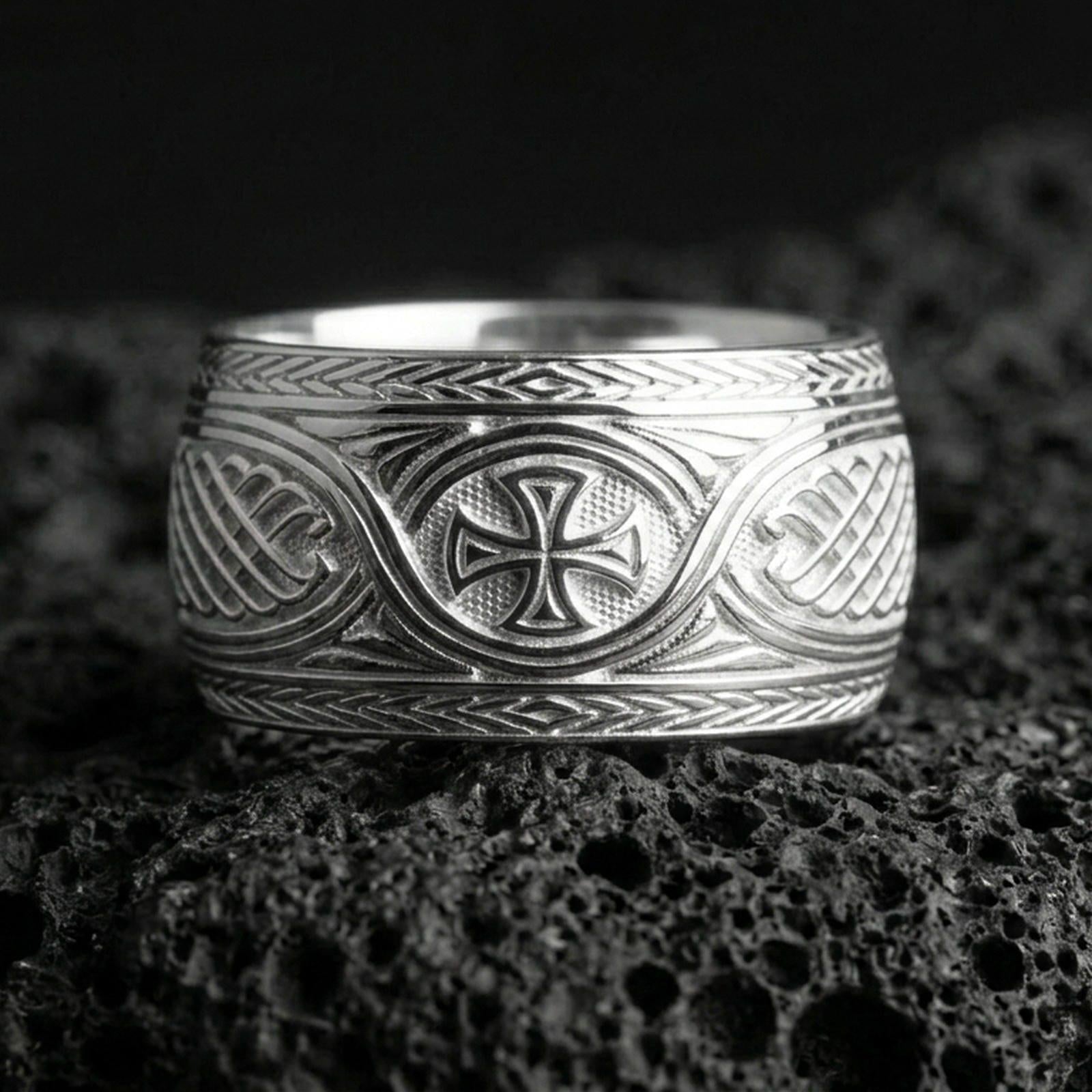 Polished silver Celtic braid band ring on volcanic rock, crisp engraved knotwork and iron cross emblem captured in sharp macro focus.