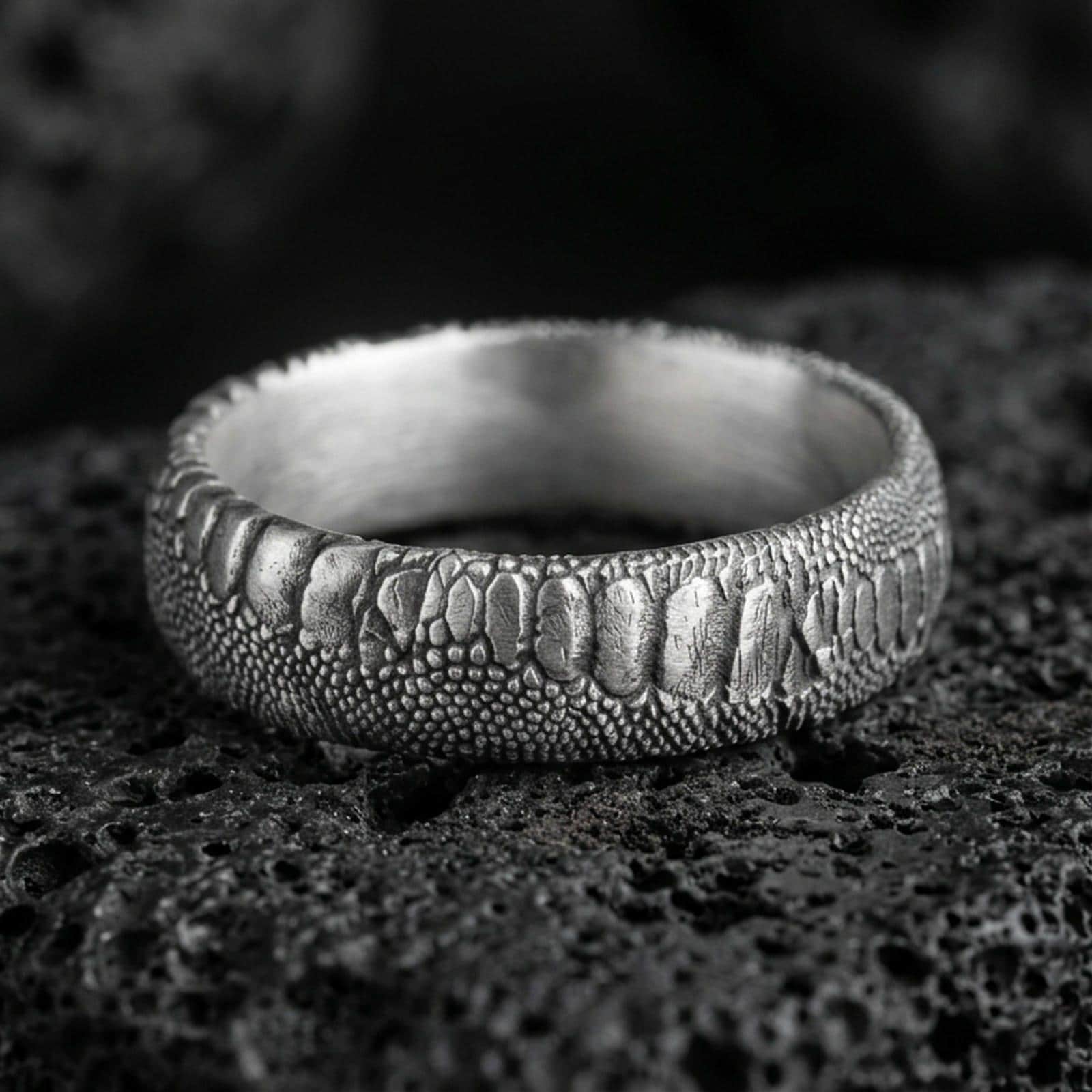 Oxidized sterling silver serpent skin band ring on black volcanic stone, scaled texture with deep shadowed recesses for a rugged gothic look.