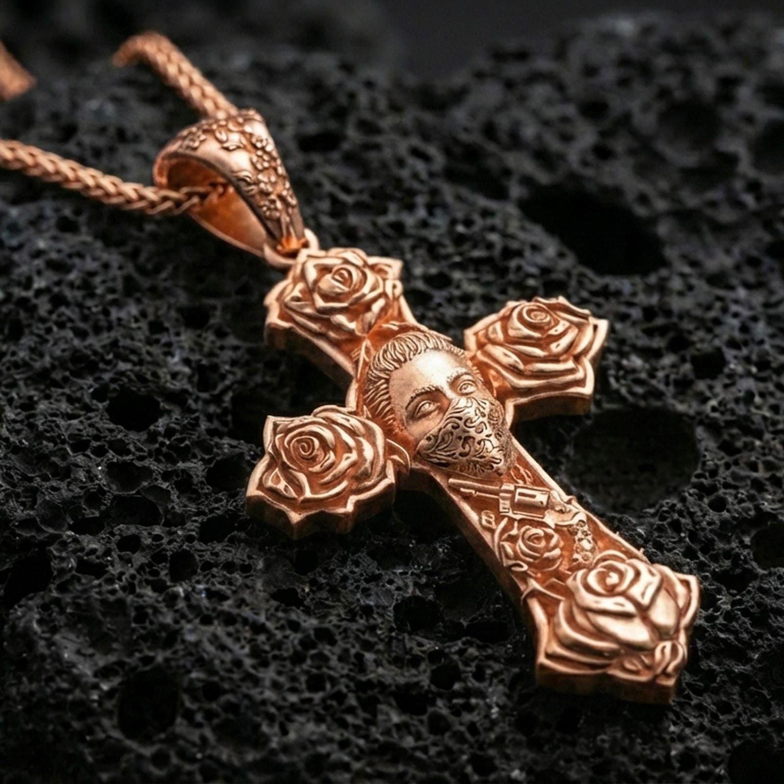 Rose gold plated cross pendant with floral cross design and engraved masked face showcased on volcanic rock with a refined close-up jewelry presentation