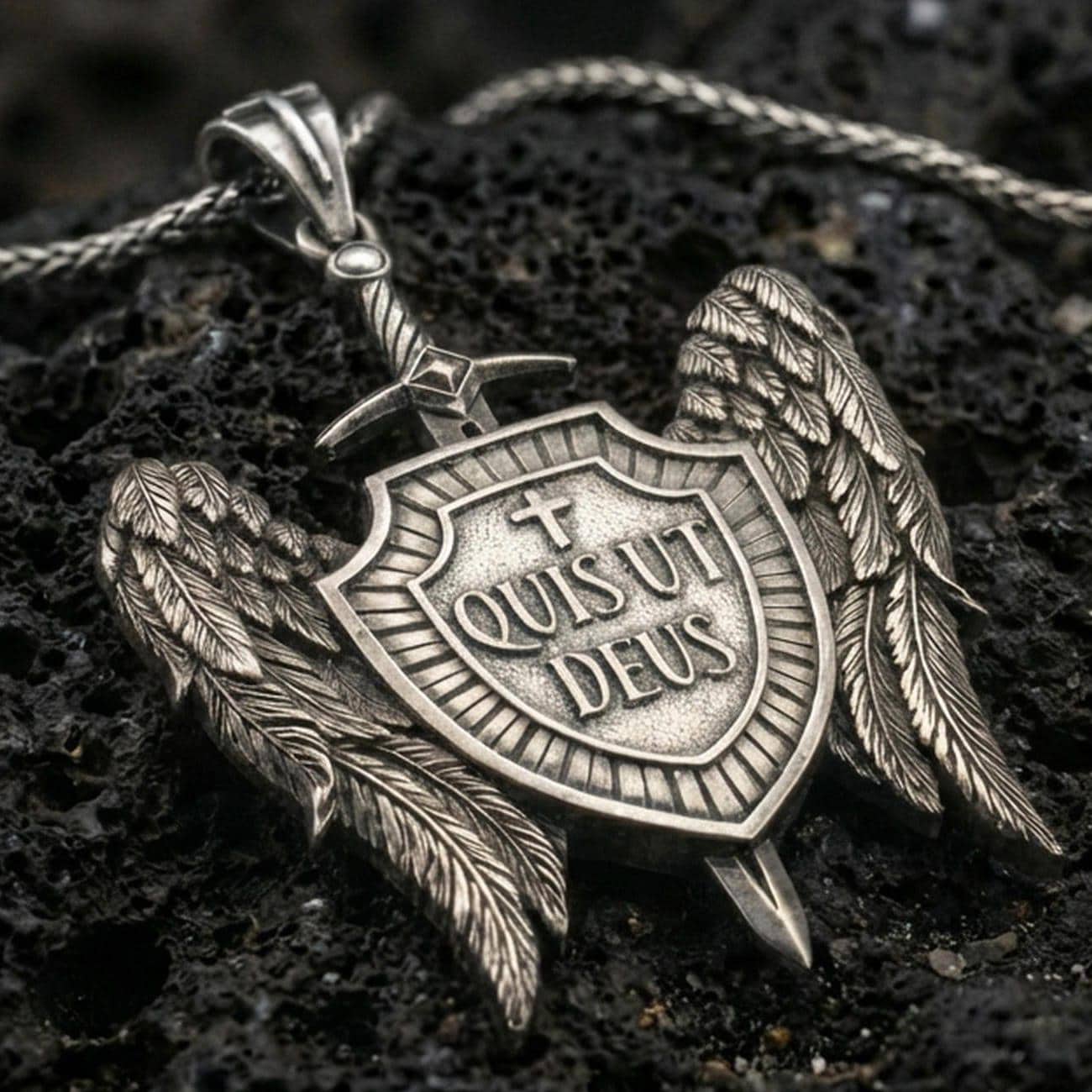 Oxidized Saint Michael inspired shield pendant with angel wings, sword detail, and Quis Ut Deus inscription displayed on dark volcanic rock in a close-up luxury jewelry shot