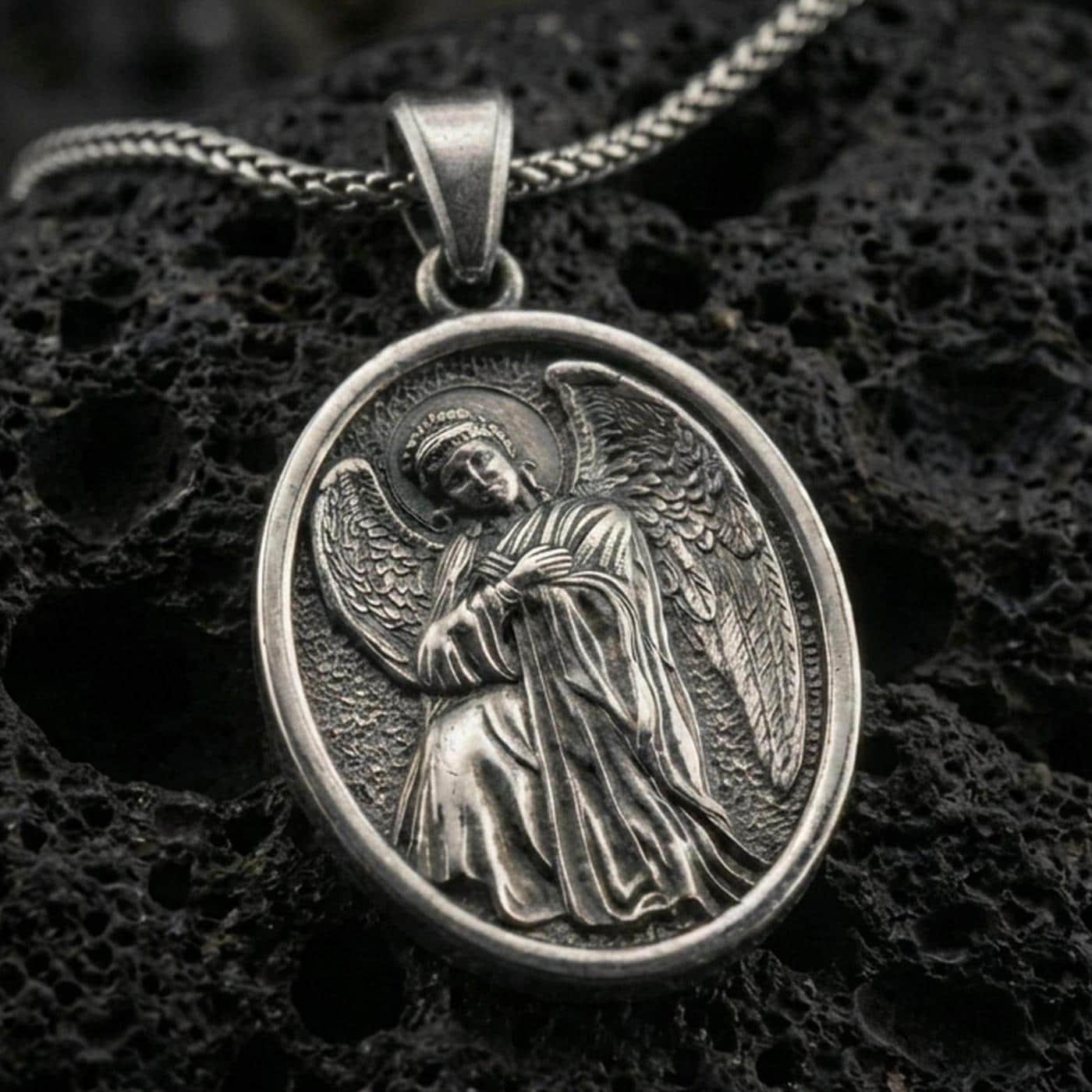 Oxidized angel pendant with oval frame and detailed winged figure displayed on dark volcanic rock in a close-up luxury jewelry composition