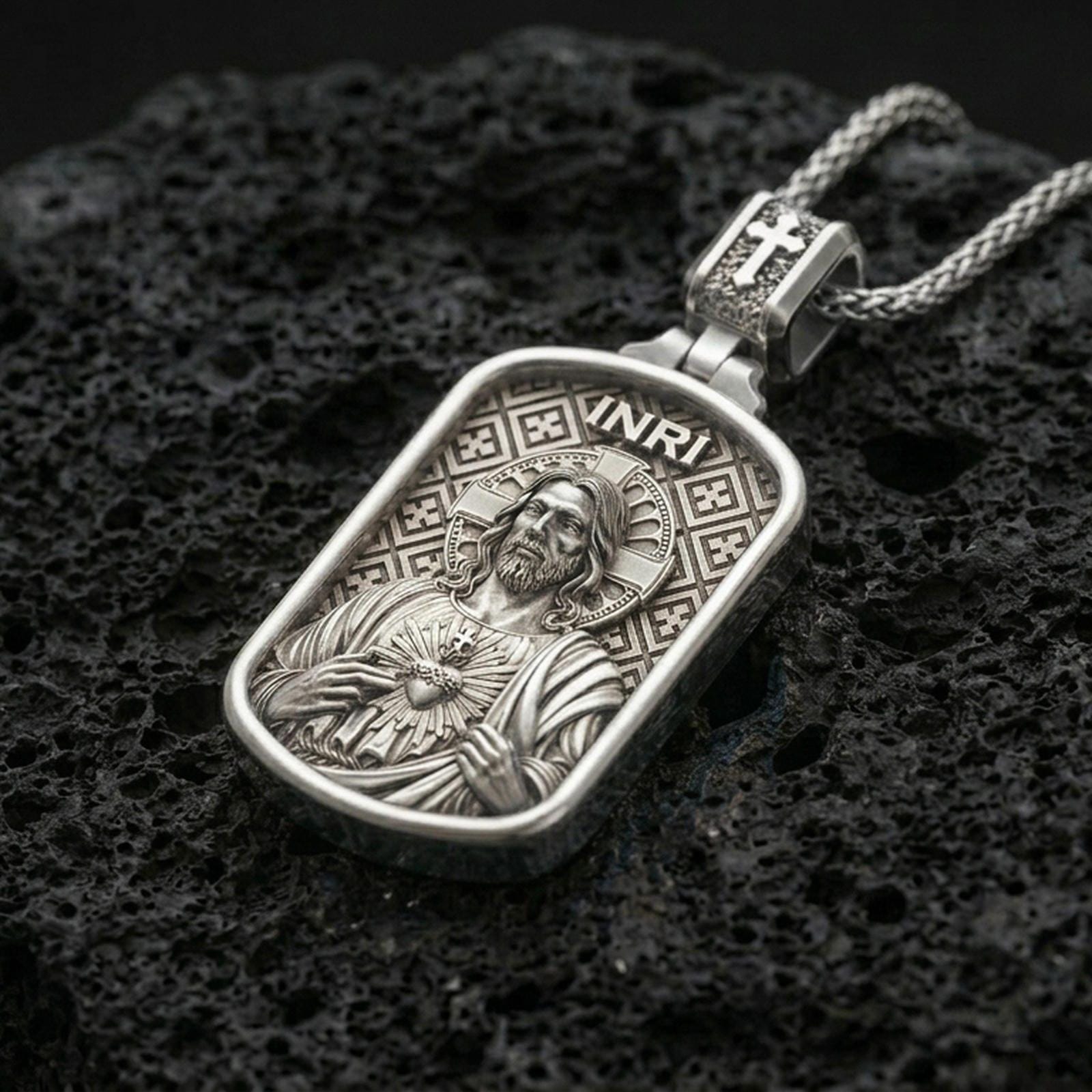 Polished silver Jesus Sacred Heart pendant with engraved halo, INRI inscription, and rounded rectangular frame placed on textured volcanic rock in a premium studio product shot