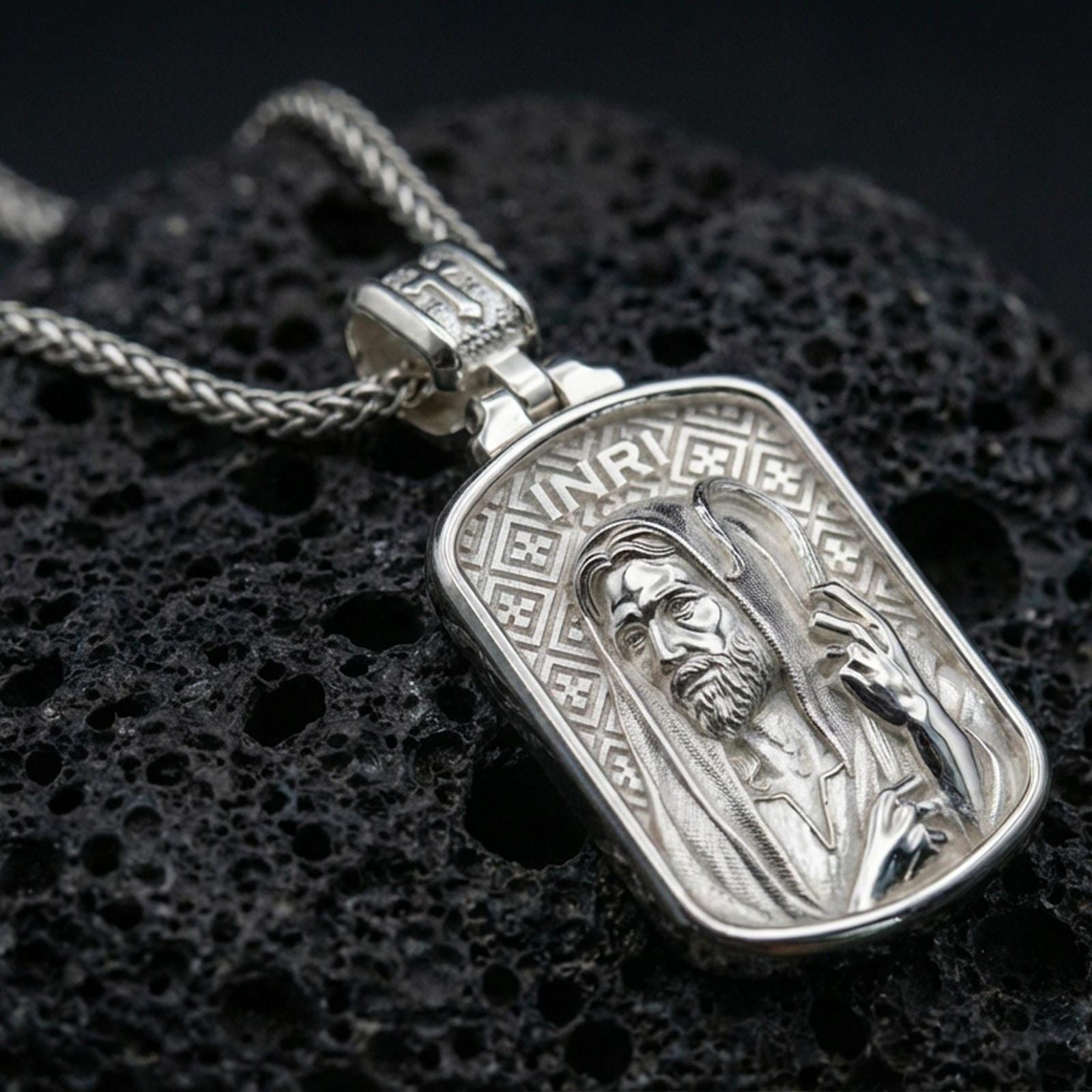 Polished silver religious pendant with Jesus portrait, INRI detail, and rounded rectangular frame placed on textured volcanic rock in a premium studio product shot