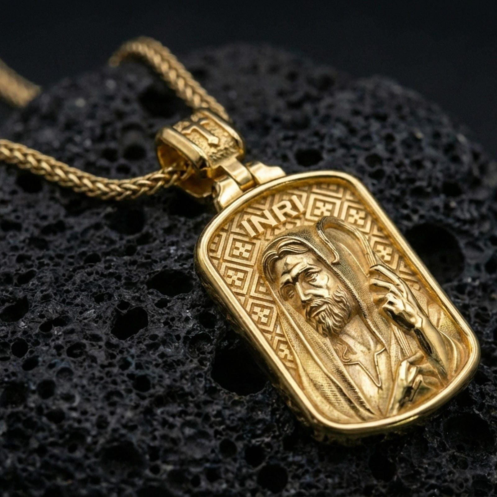 Gold plated Christian pendant with hooded Jesus figure, staff detail, and INRI lettering showcased on black volcanic rock in a refined macro jewelry image