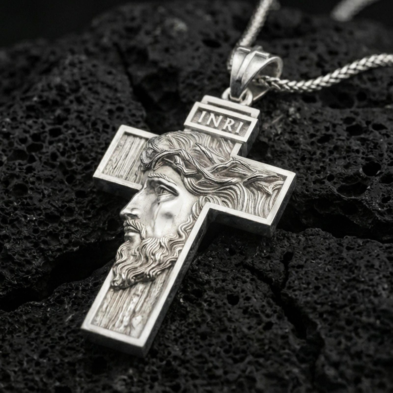 Polished silver Christian cross pendant with sculpted Jesus face, wood-textured cross relief, and INRI inscription placed on textured volcanic rock in a premium studio product shot