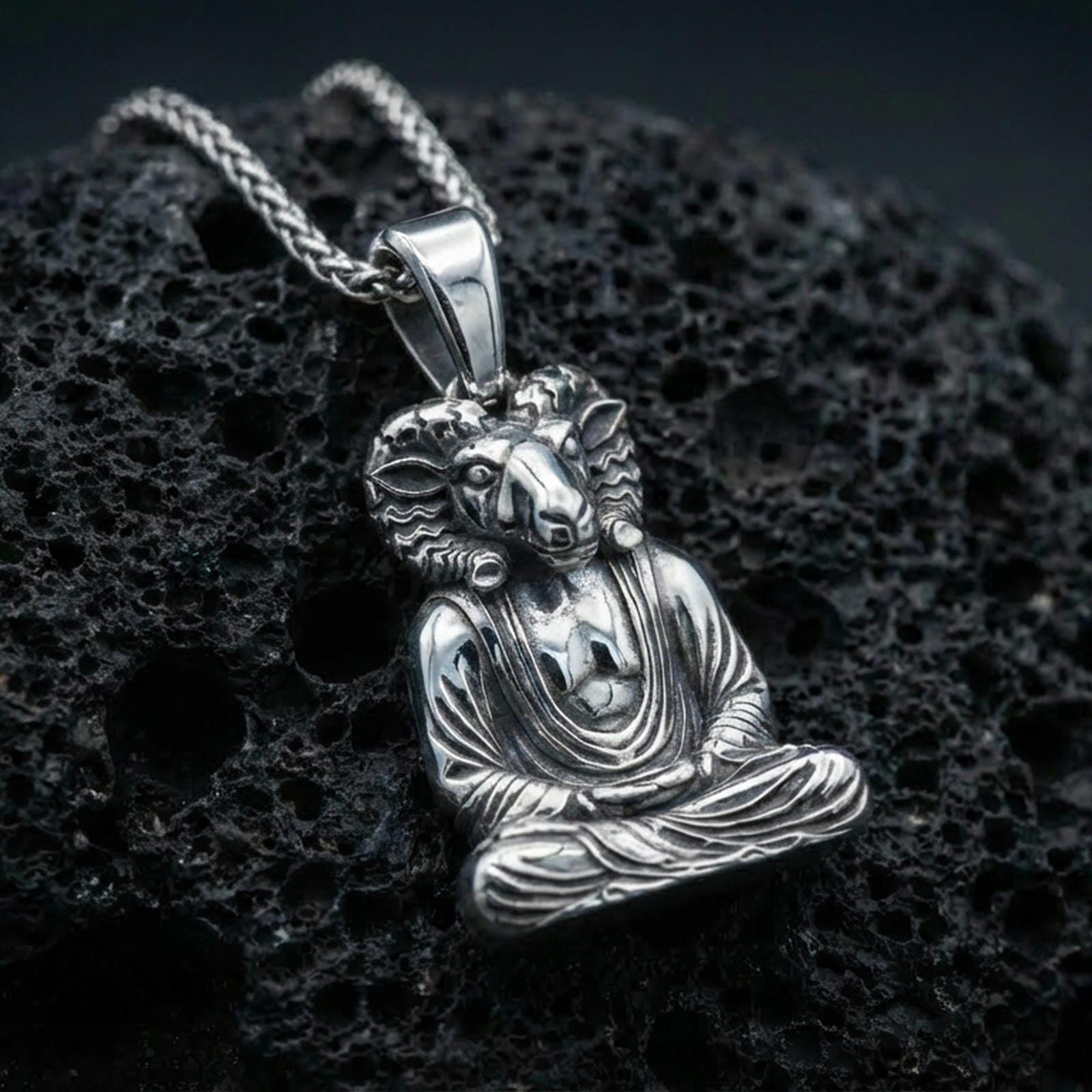 Sterling silver Egyptian god pendant in a polished silver finish, Khnum ram-head meditation figure with sculpted facial details and draped garment, close-up on volcanic stone