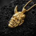Load image into Gallery viewer, Sterling silver oni mask pendant with a gold plated finish, Japanese demon mask with horns and sharp teeth, close-up on volcanic rock
