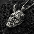 Load image into Gallery viewer, Sterling silver oni mask pendant with an oxidized finish, Japanese demon face with horns and fangs in high relief, close-up on black volcanic rock
