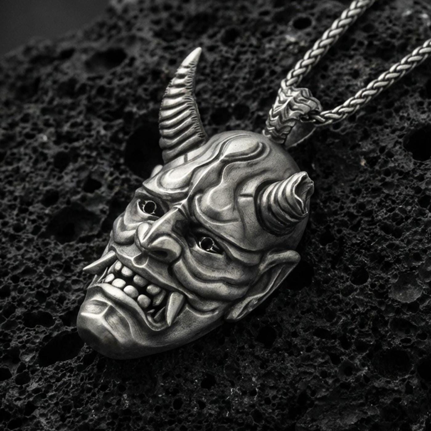 Sterling silver oni mask pendant with an oxidized finish, Japanese demon face with horns and fangs in high relief, close-up on black volcanic rock
