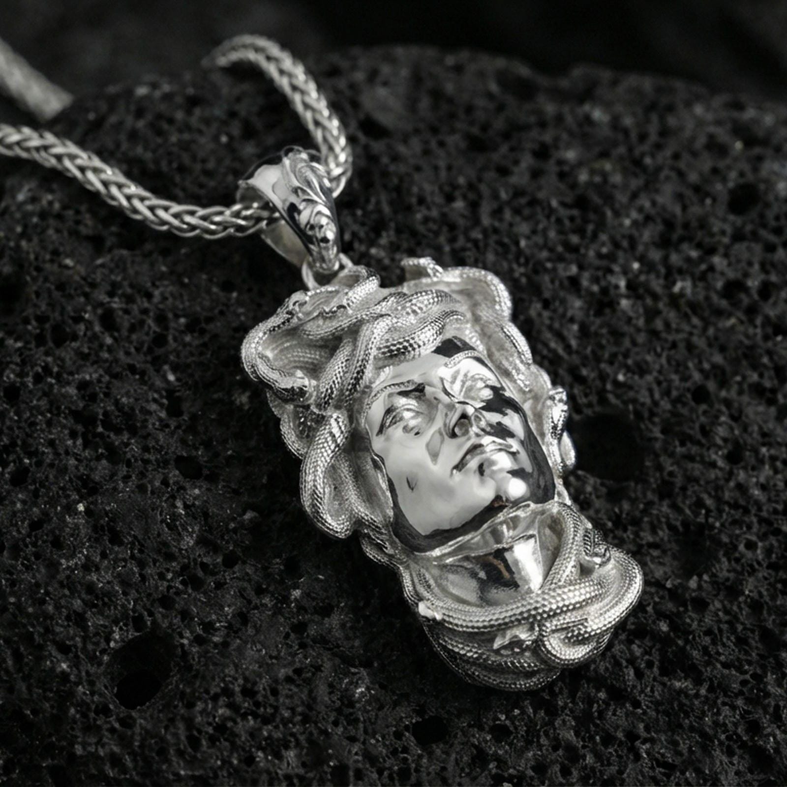 Polished silver Medusa head pendant necklace on textured volcanic rock, crisp close-up product shot highlighting the sculpted face and coiled serpent details.