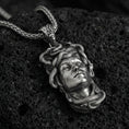 Load image into Gallery viewer, Oxidized Medusa head pendant necklace on textured volcanic rock, close-up centered composition with detailed snake relief and dramatic studio lighting.
