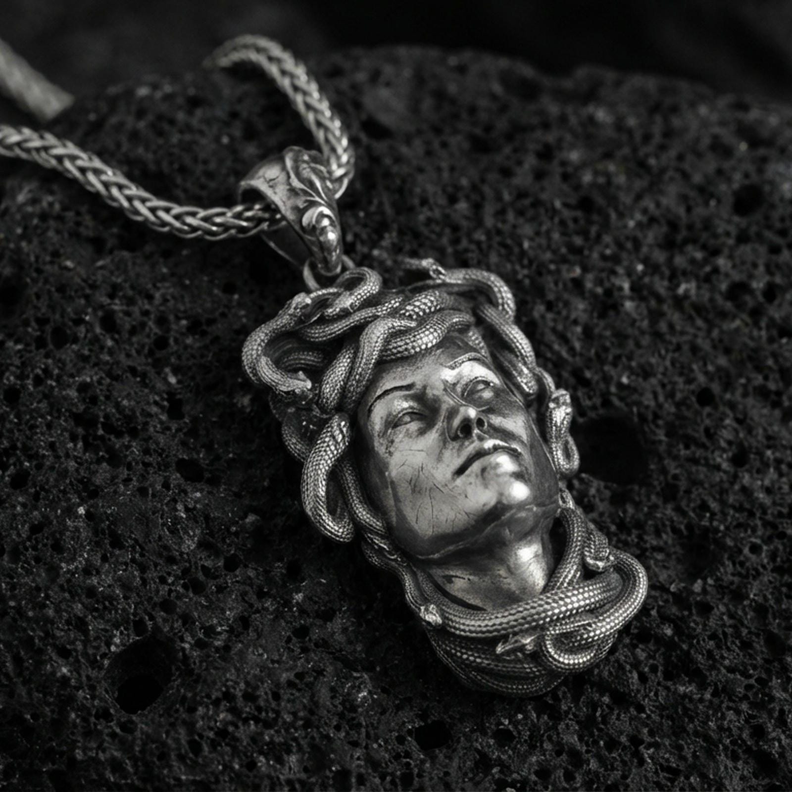 Oxidized Medusa head pendant necklace on textured volcanic rock, close-up centered composition with detailed snake relief and dramatic studio lighting.