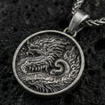 Load image into Gallery viewer, Oxidized round dragon coin pendant necklace on textured volcanic rock, centered close-up highlighting the engraved scales, flowing mane, and deep antique contrast.
