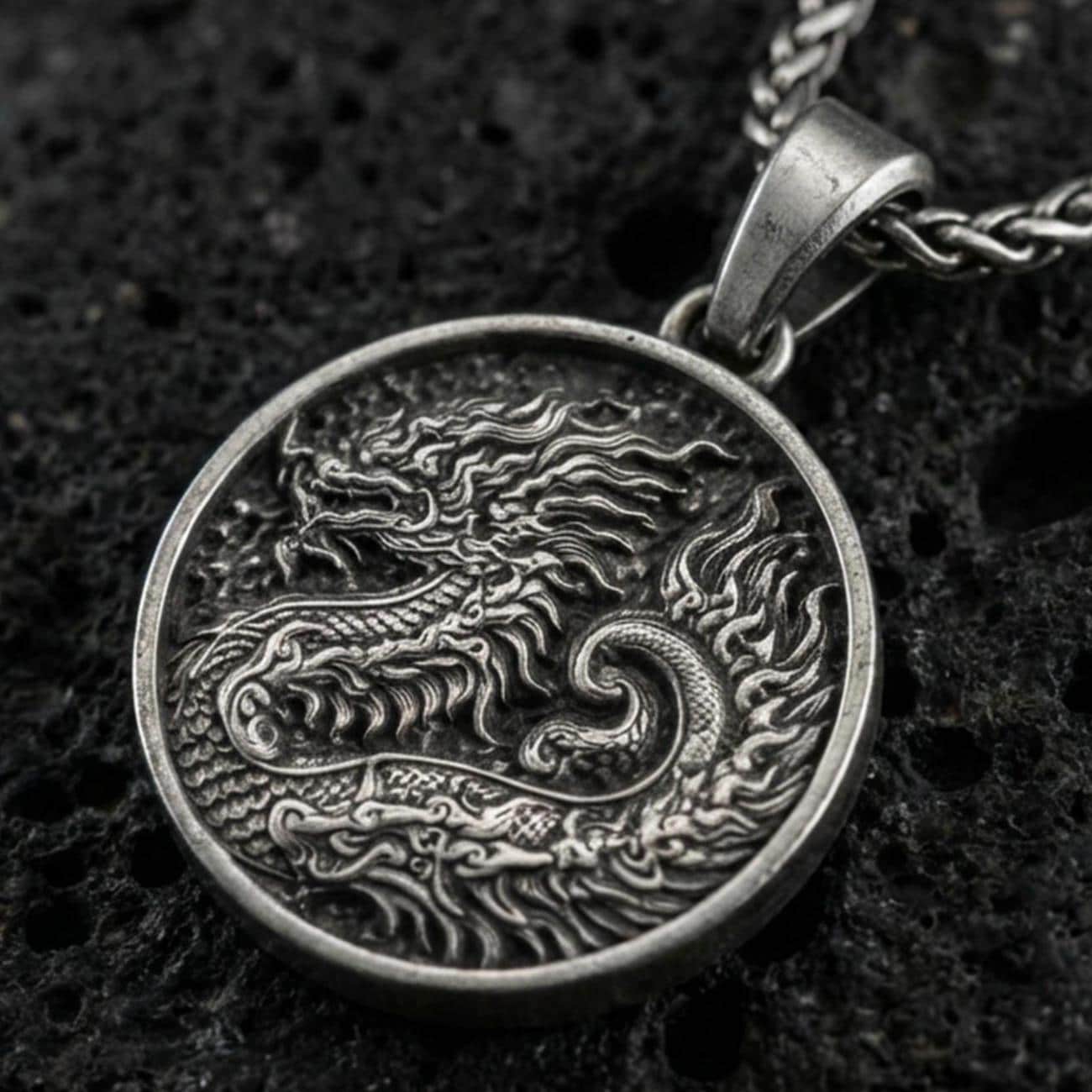 Oxidized round dragon coin pendant necklace on textured volcanic rock, centered close-up highlighting the engraved scales, flowing mane, and deep antique contrast.