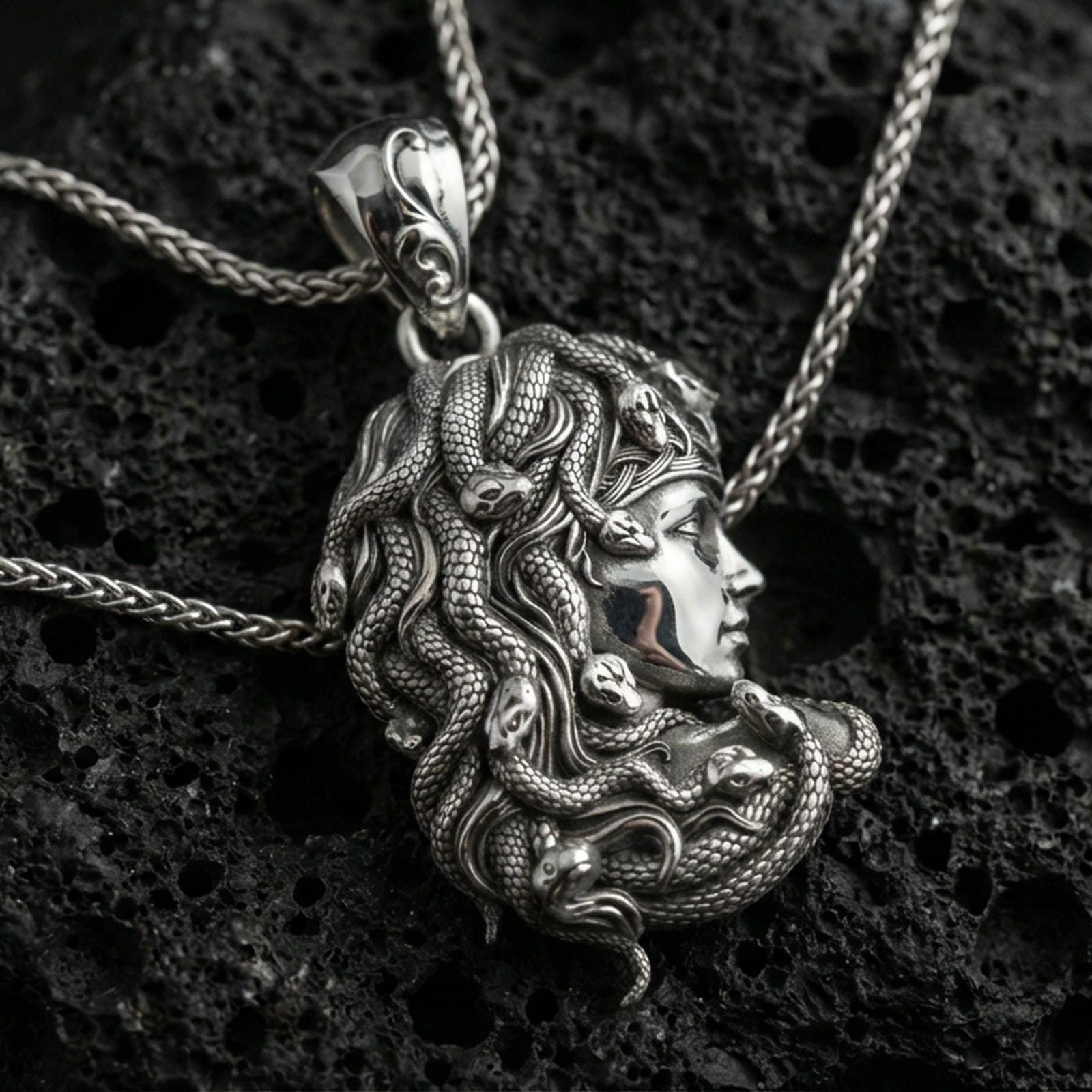 Polished silver Medusa profile pendant necklace on textured volcanic rock, crisp macro product photo emphasizing smooth facial contours, sharp serpent detailing, and clean reflective edges.