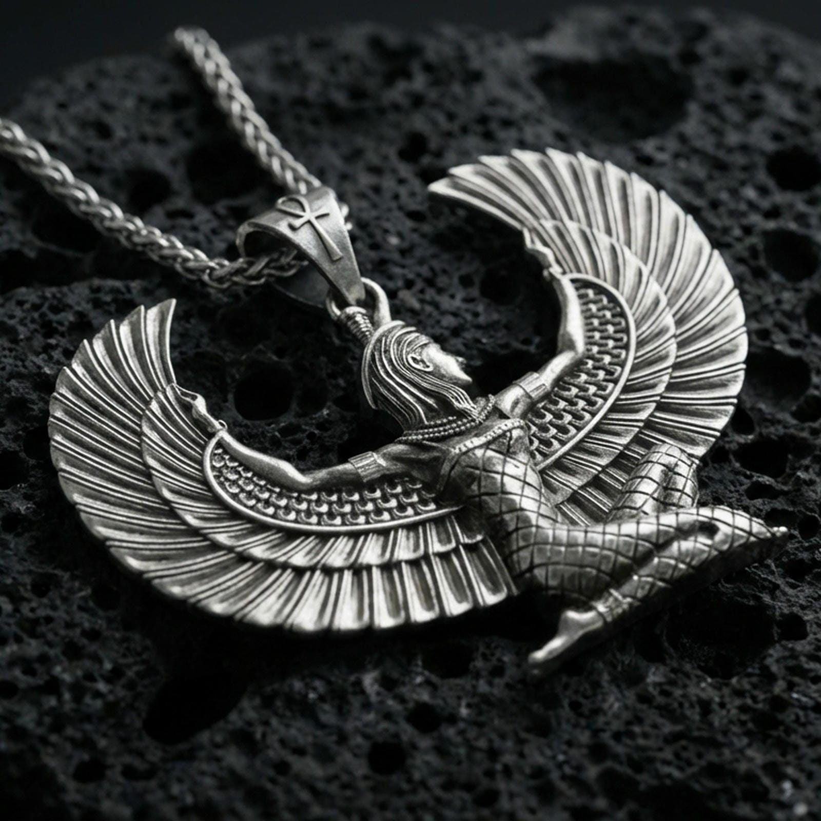 Oxidized winged goddess Isis pendant necklace on textured volcanic rock, centered close-up highlighting the spread wings, feather lines, and deep antique shadowed contrast.