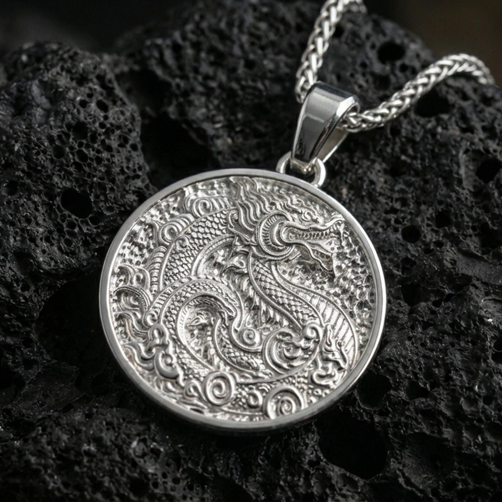Polished silver dragon medallion pendant necklace on textured volcanic rock, crisp macro product photo emphasizing sharp scale texture, clean edges, and detailed engraved relief.