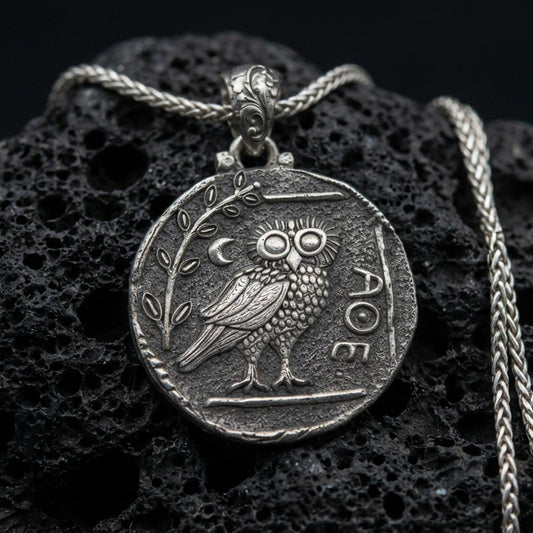 Oxidized owl coin pendant necklace on textured volcanic rock, centered close-up highlighting the ancient-style owl relief, moon detail, and deep antique contrast on the matte background.