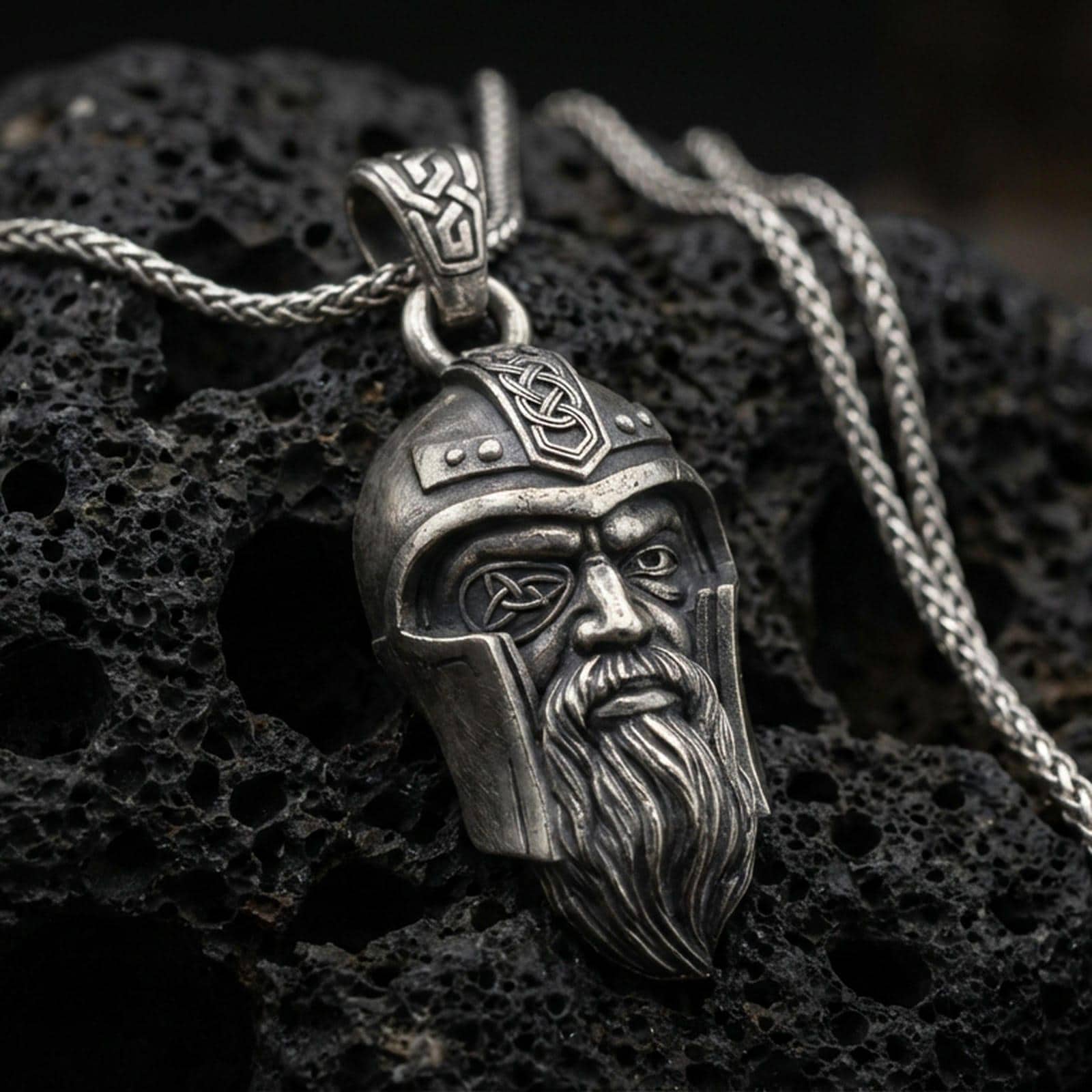 Oxidized Odin pendant necklace on textured volcanic rock, centered close-up highlighting the one-eyed helmeted face, braided beard, and deep antique shadowed contrast.